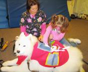woman in a nurses outfit sitting with and looking on as a young girl in a surgical mask pets a white dog (looks like a samoyed)