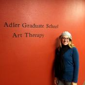 Erin standing against a red wall with the words "Adler Graduate School Art Therapy" on it. 