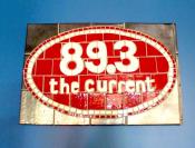 a photo of The Current logo, which says, "89.3 the current logo". The text is white on a horizontal red oval with a white and red border on the oval. 